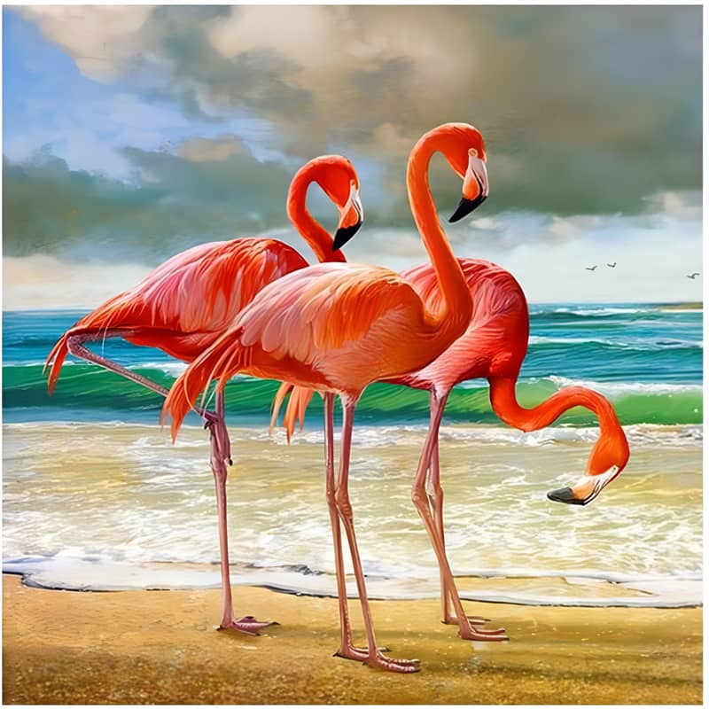Three vibrant flamingos standing on a sandy beach with ocean waves and a cloudy sky in the background.