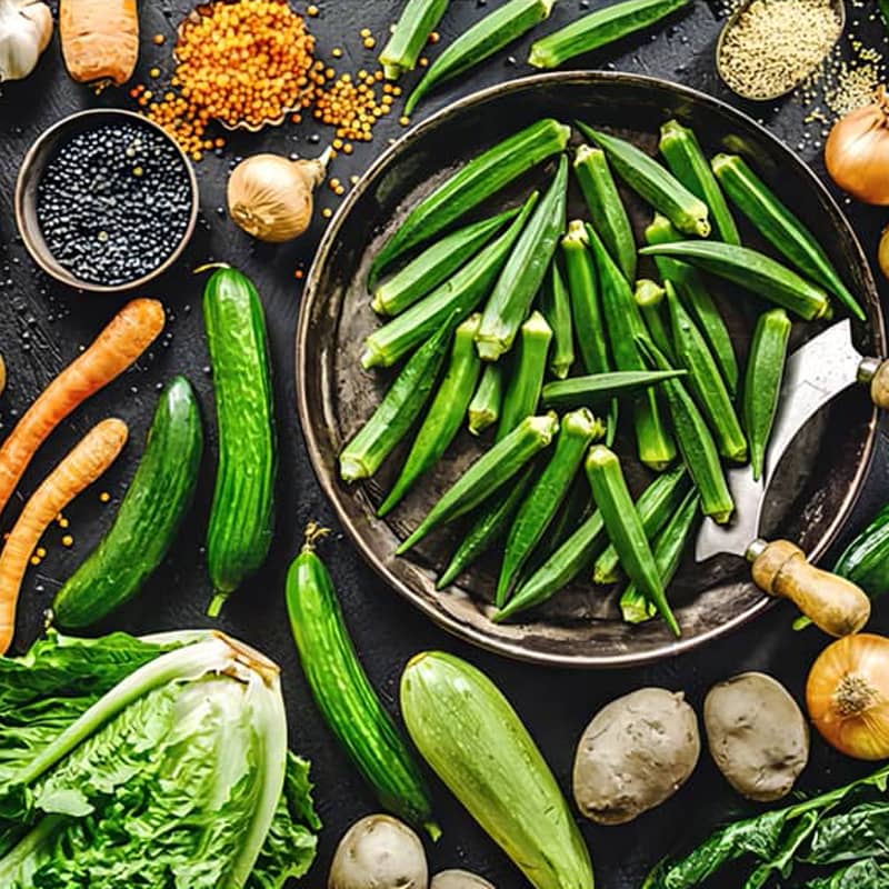 Assorted fresh vegetables including okra, lettuce, carrots, cucumbers, onions, and potatoes displayed on a dark background.