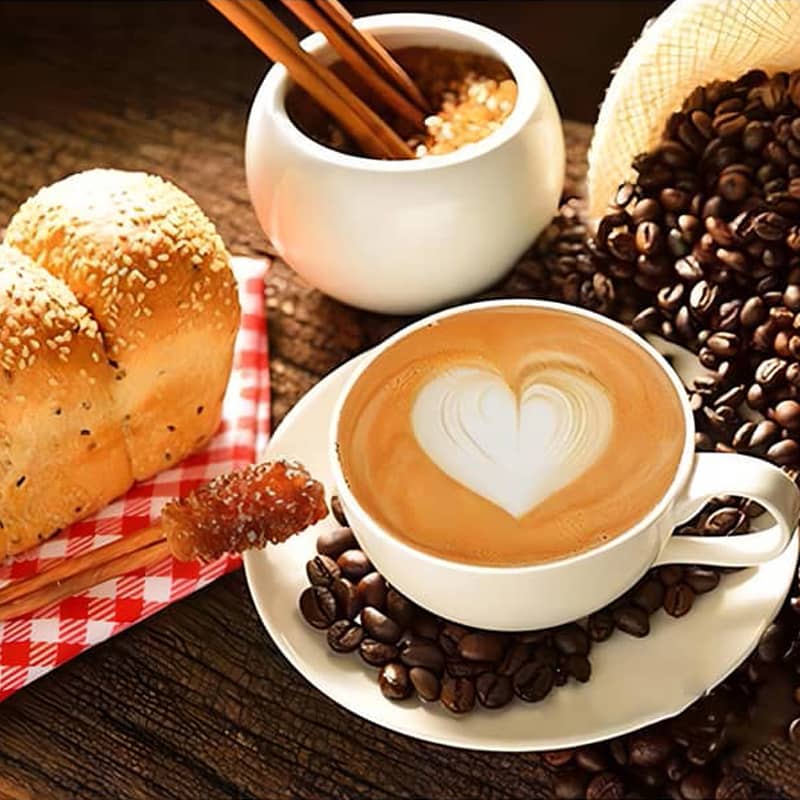 Cappuccino with heart latte art surrounded by coffee beans, sesame bread, and sugar sticks on a rustic wooden table.