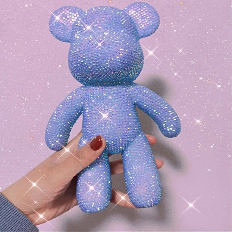 Hand holding a sparkling purple diamond painting Glitter Bear against a light pink background